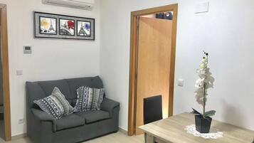 Una sala de estar con un sofá gris, cojines estampados, una mesa de comedor de madera con una orquídea blanca y una puerta con un espejo.