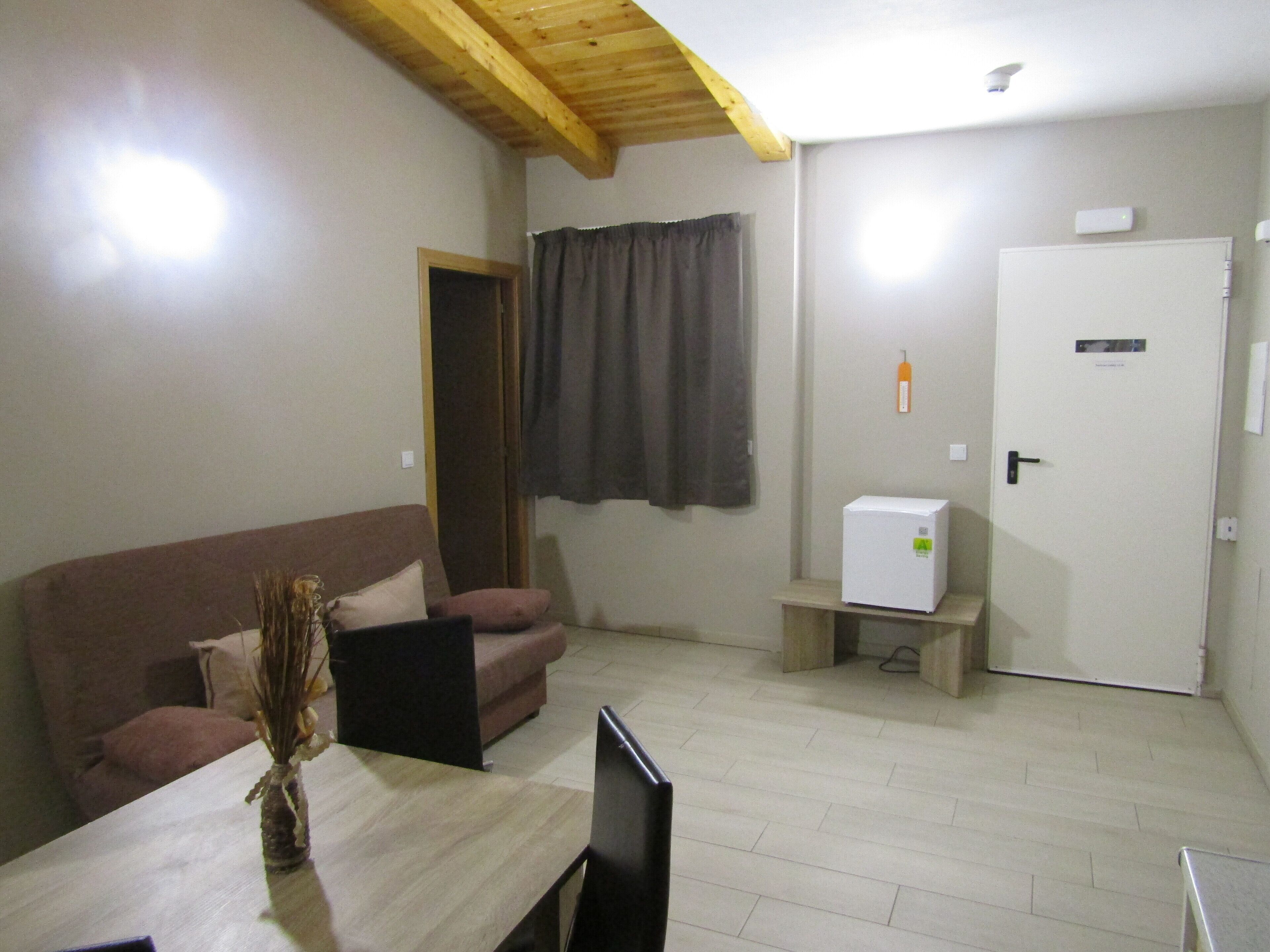 Una habitació amb sostre de fusta, un sofà marró, una taula petita amb un ram i un frigorífic blanc, i una porta amb un cartell.