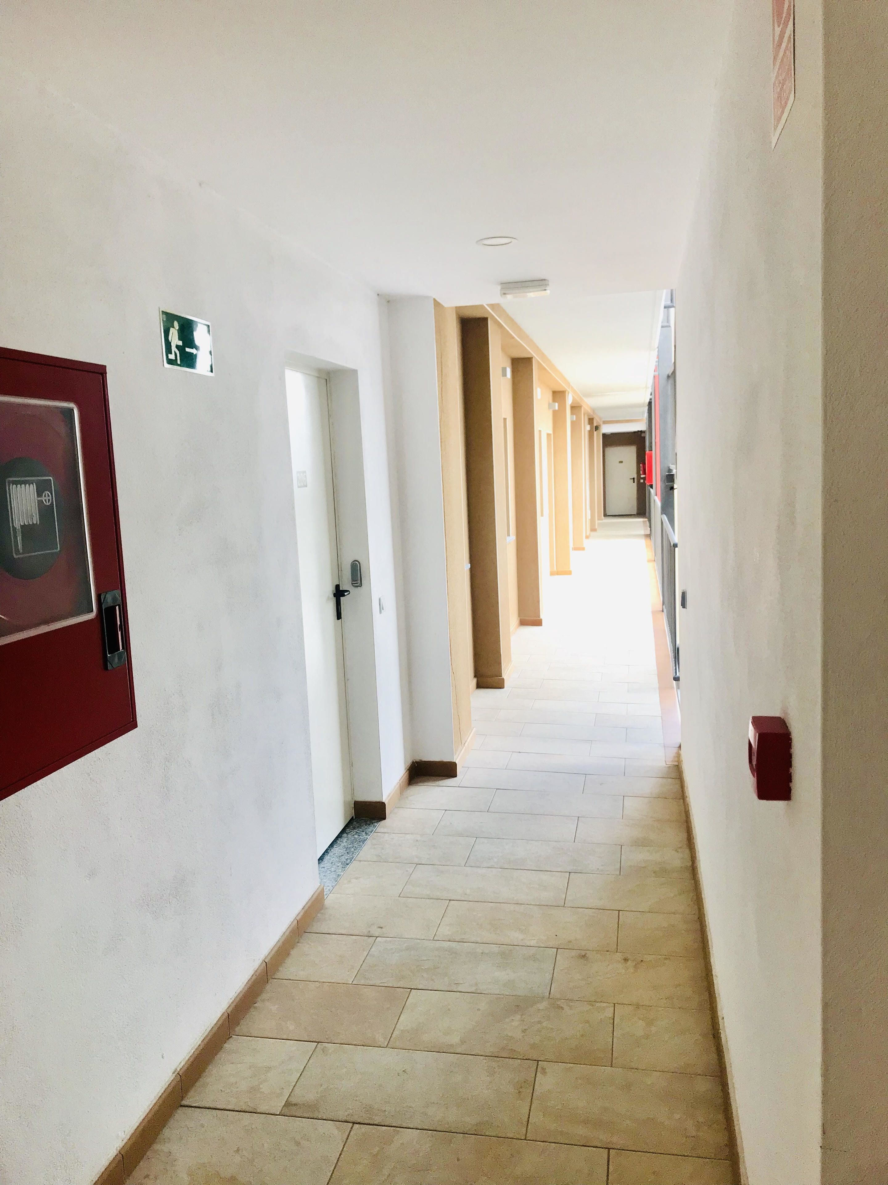 Un pasillo largo y estrecho con puertas a ambos lados, un detector de incendios a la izquierda y un extintor a la derecha.