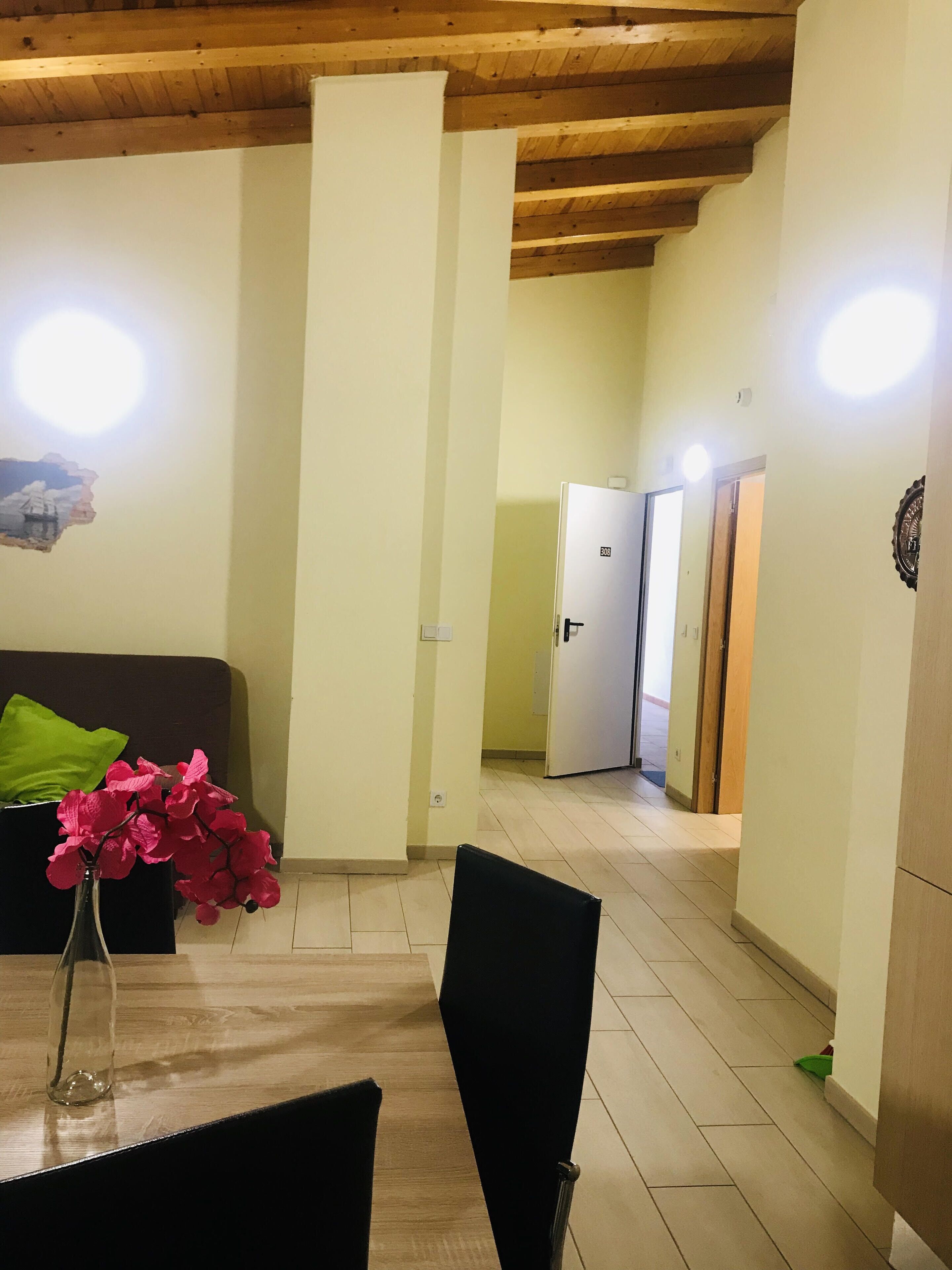 A room with a wooden ceiling, a sofa with green cushions, a vase with pink flowers, and a door leading to another room.