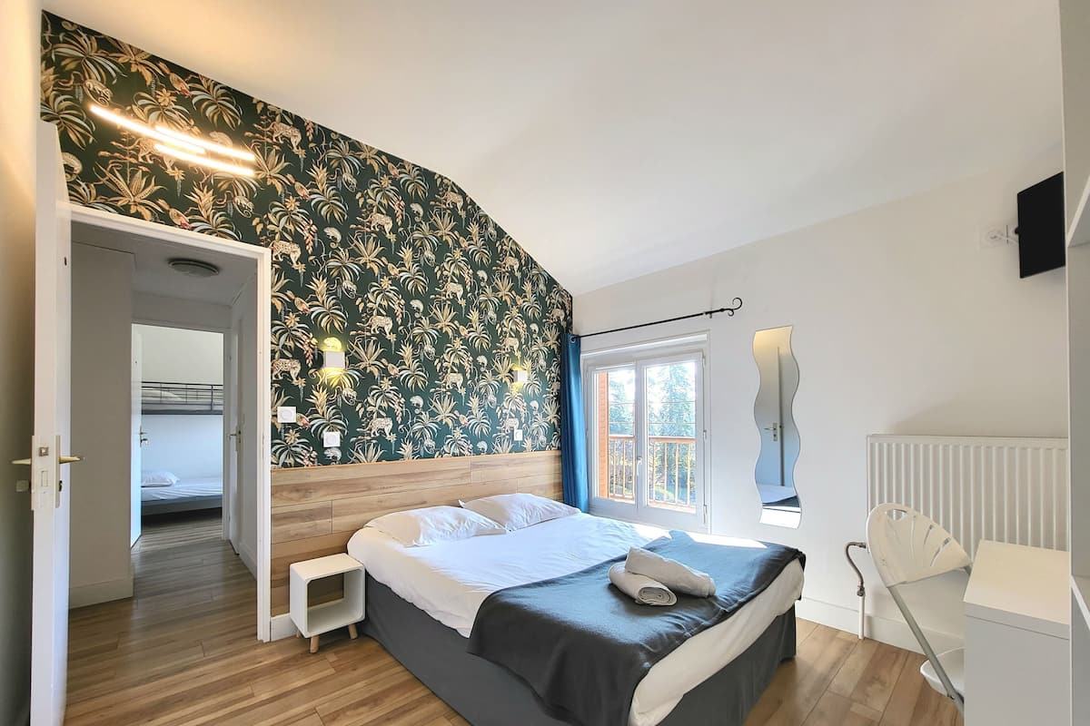 Een slaapkamer met een bed, nachtkastje, spiegel en raam met uitzicht op bomen.