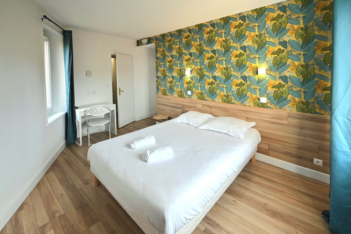 Een slaapkamer met een houten hoofdeinde, een bed met witte lakens, een bureau met stoel en een wand met een bladmotief.