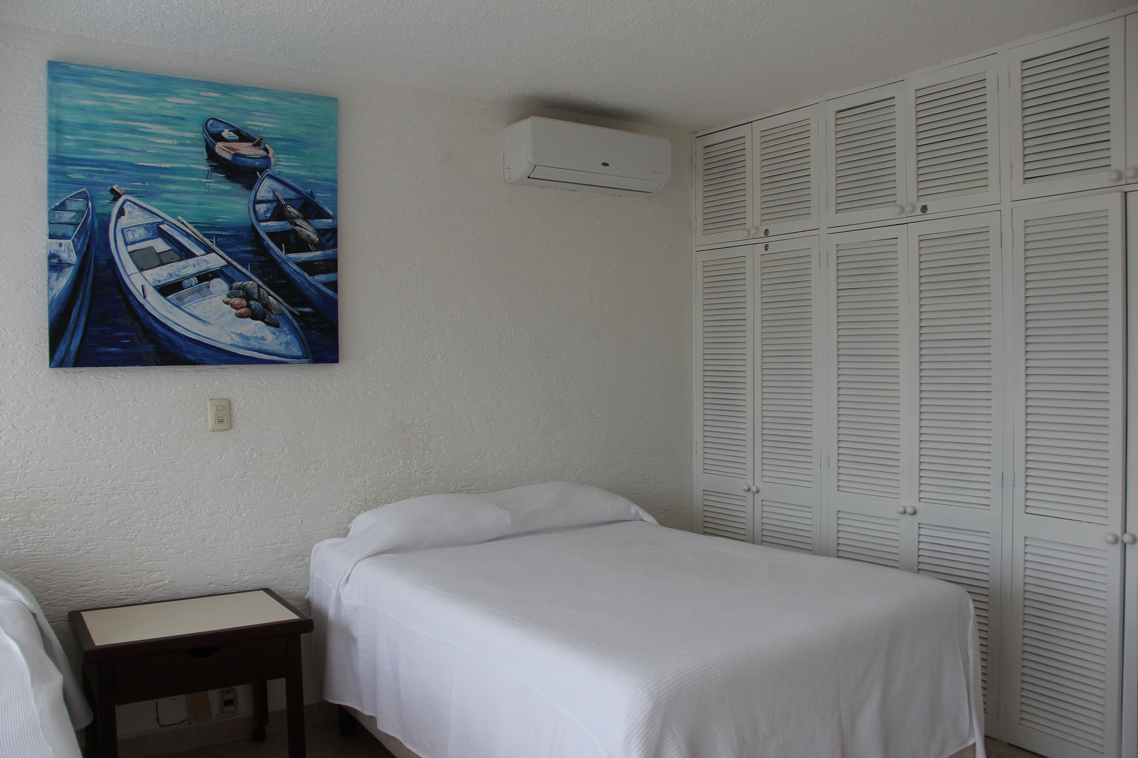 Een slaapkamer met een bed, een nachtkastje, een schilderij van boten, een witte airconditioning en een set witte louvre-kasten.