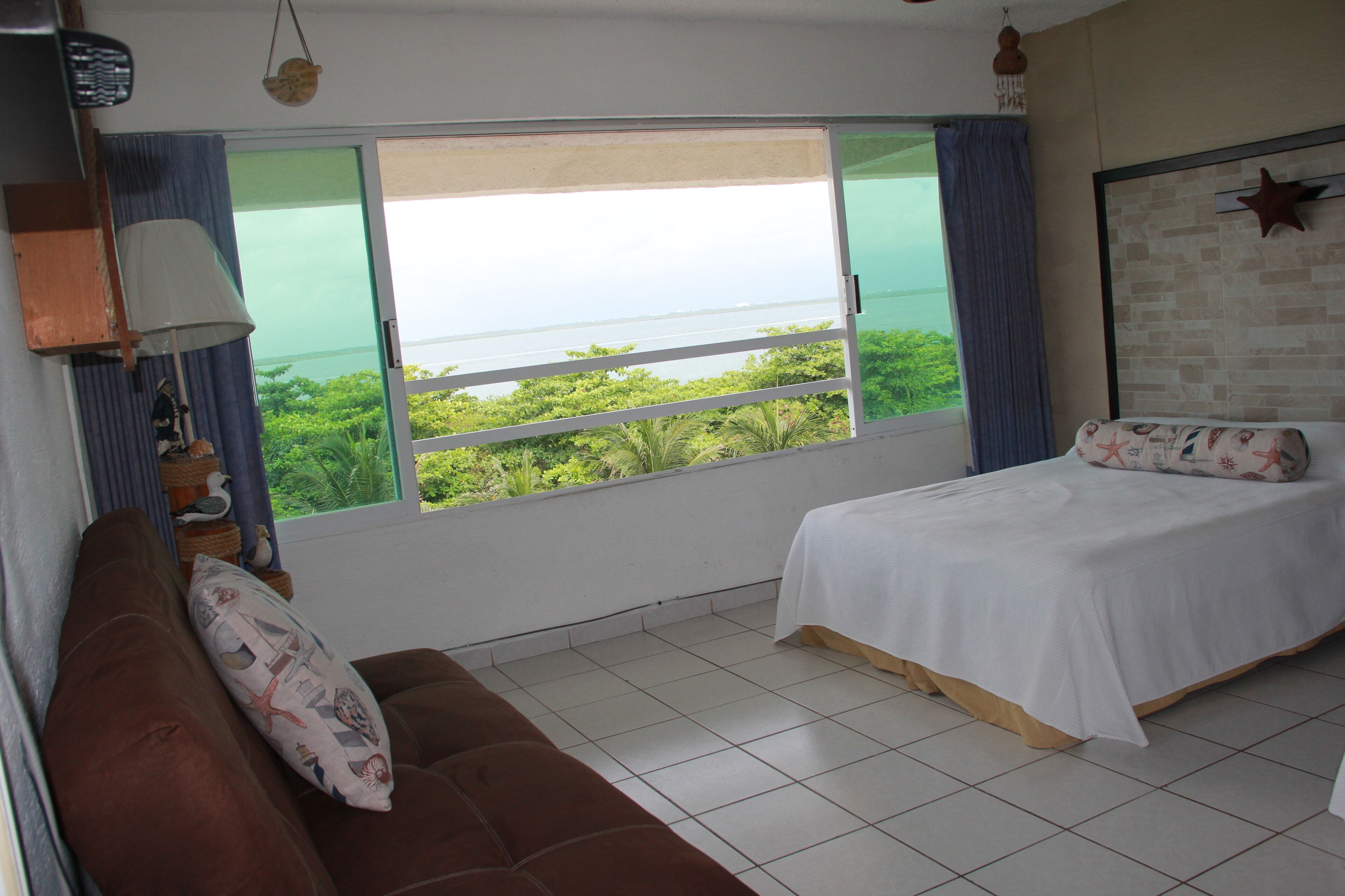 A bedroom with a large window, a bed, a sofa, and a lamp.