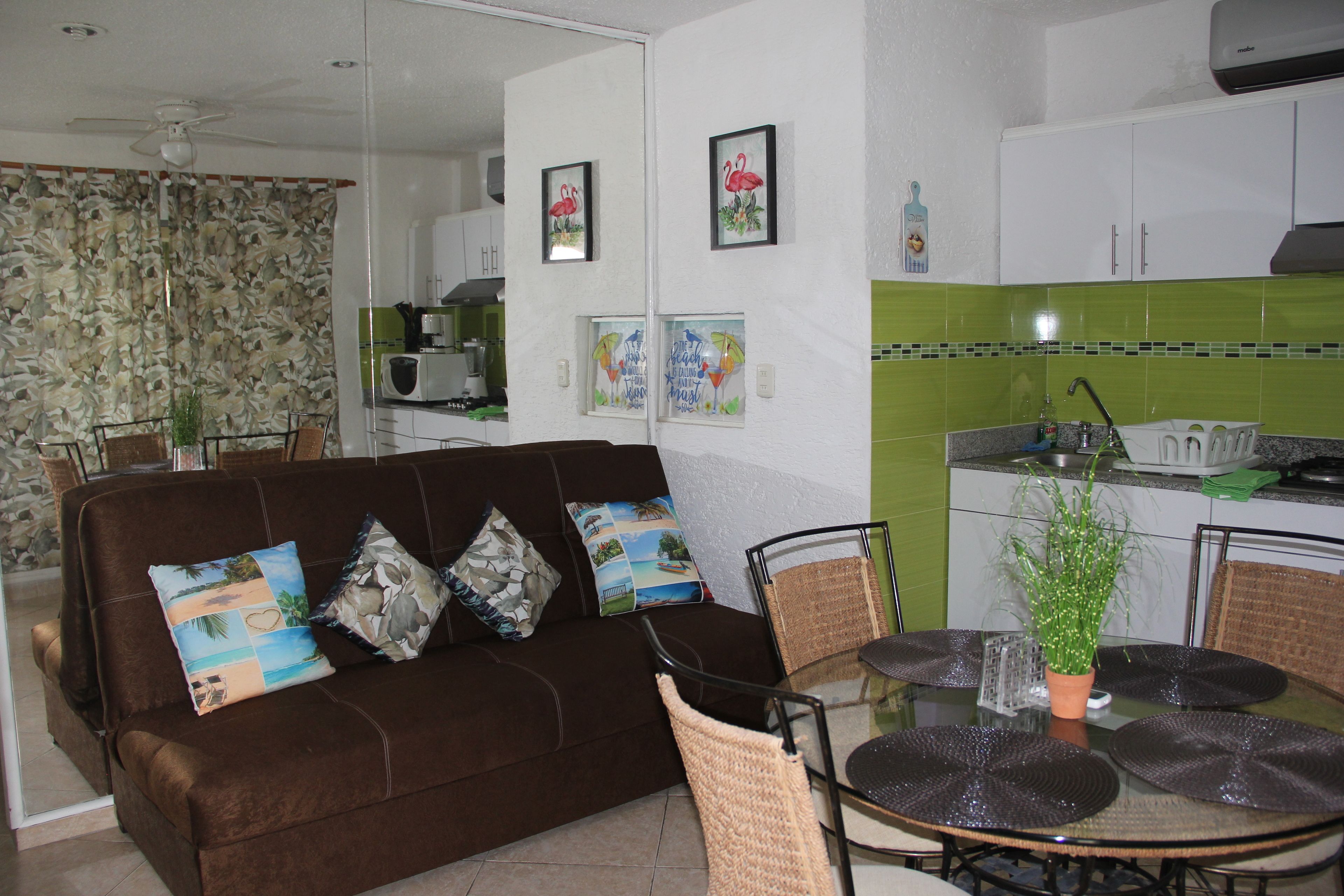 A living room with a brown sofa, two chairs, a glass table, and a kitchen area with a microwave and green tiles.