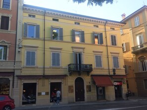 Um edifício de vários andares com fachada amarela, várias janelas com persianas e entrada no andar térreo com toldo vermelho.