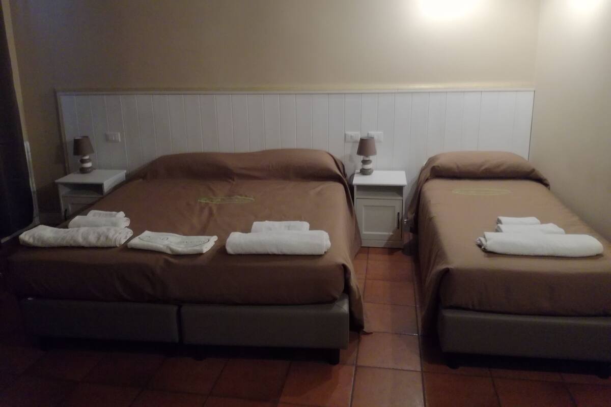 Ein Hotelzimmer mit zwei Einzelbetten, jeweils mit Kissen und einem Handtuch. Dazwischen befindet sich ein Nachttisch mit einer Lampe.