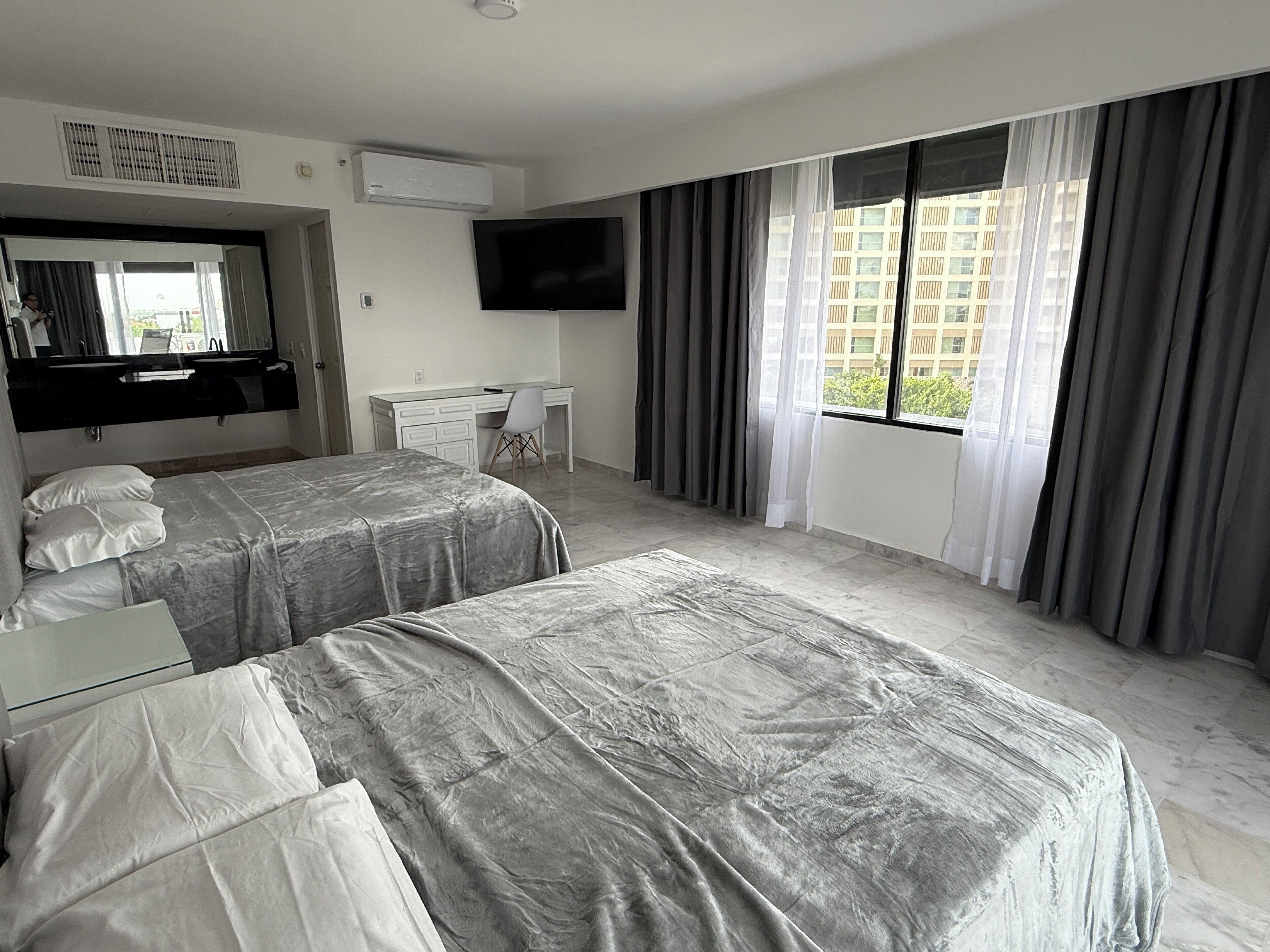 A hotel room with two beds, a desk, a TV, and a window with curtains.