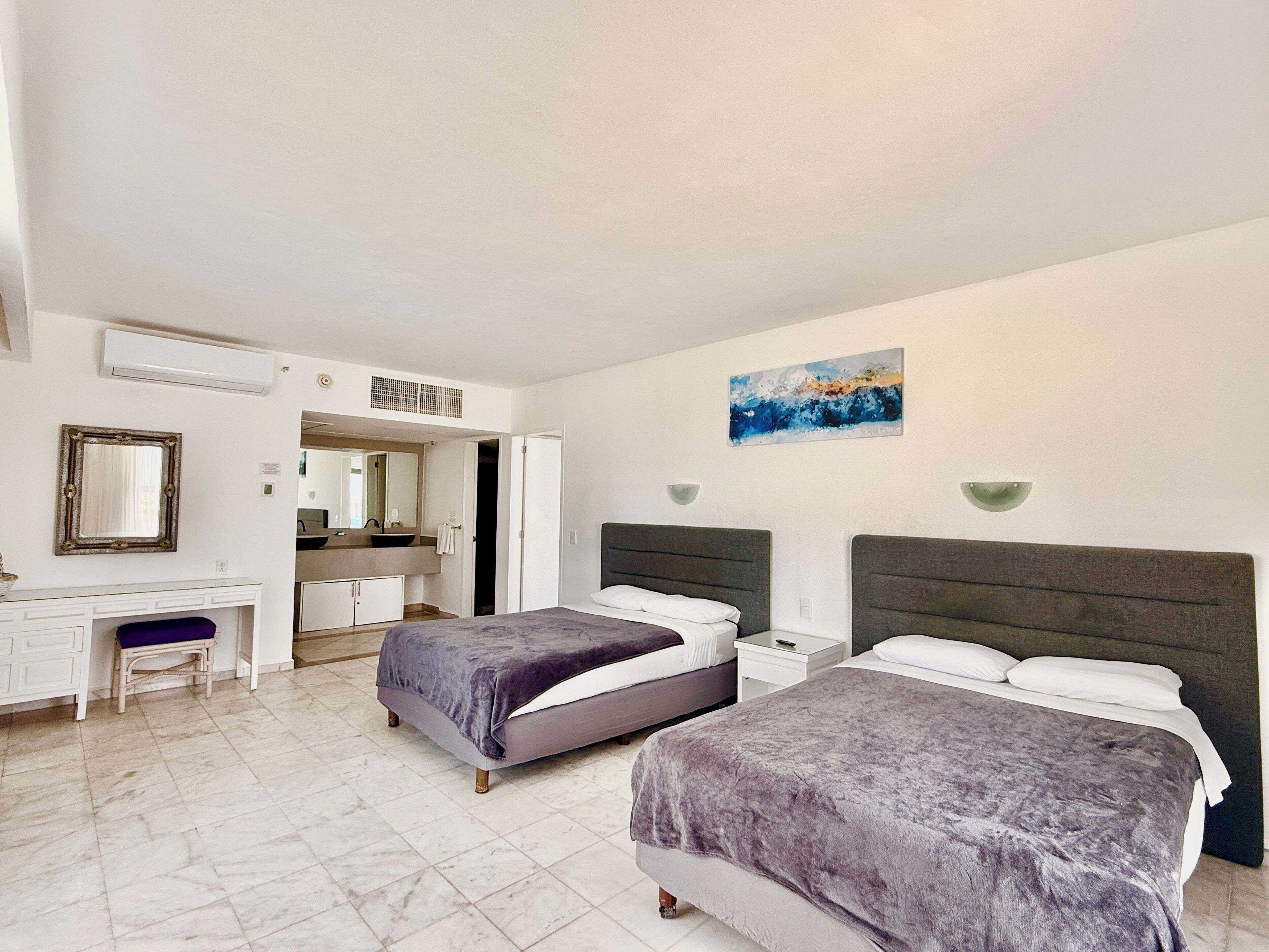 A hotel room with two beds, a desk, a chair, a vanity with a mirror, and a painting on the wall.