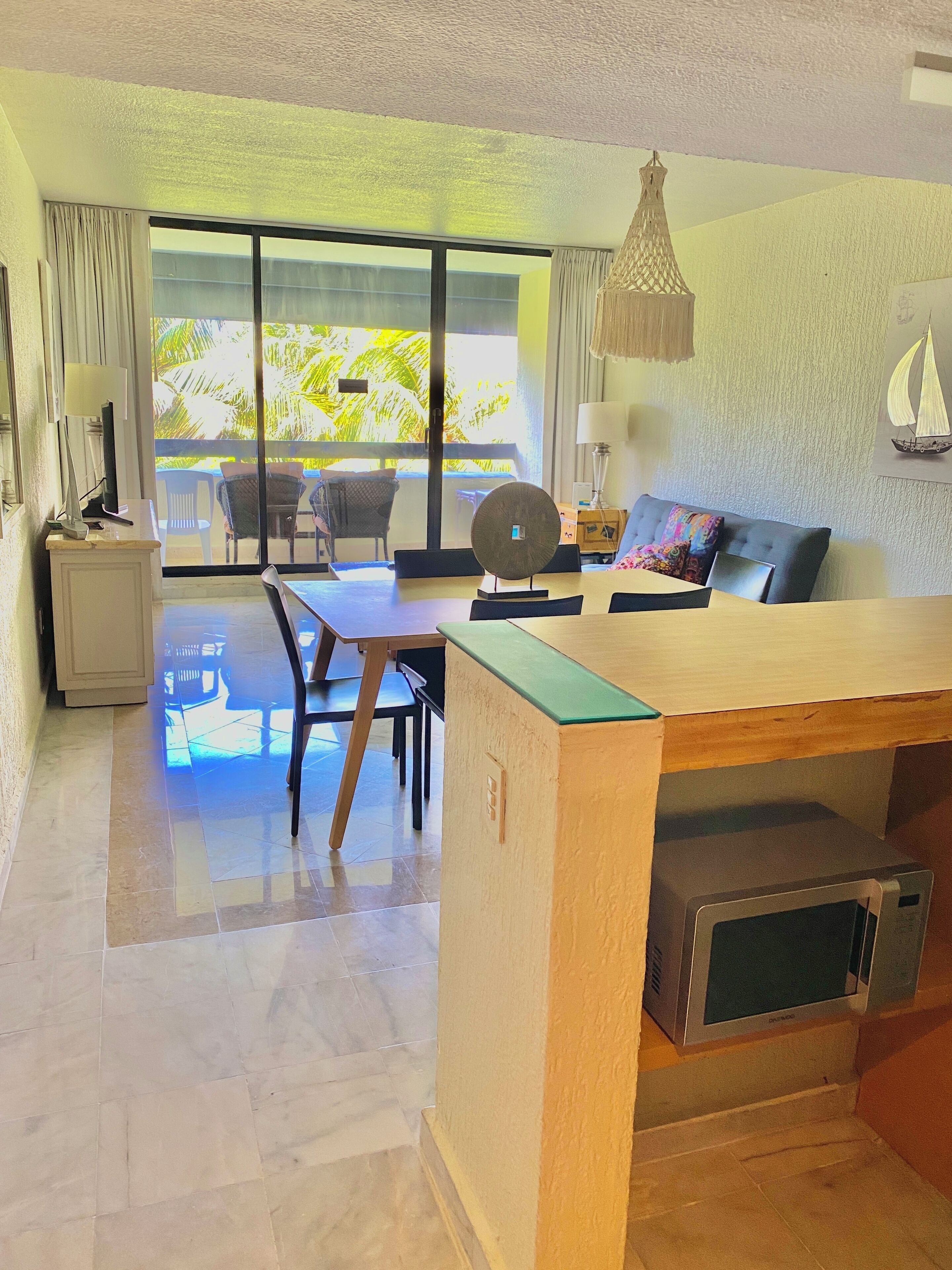 A modern living room with a dining area, a large sliding glass door leading to a balcony, a microwave, and a small table with a lamp.