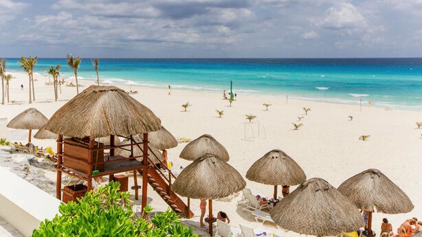 Een strand met rieten parasols, houten strandhutjes en witte ligstoelen.