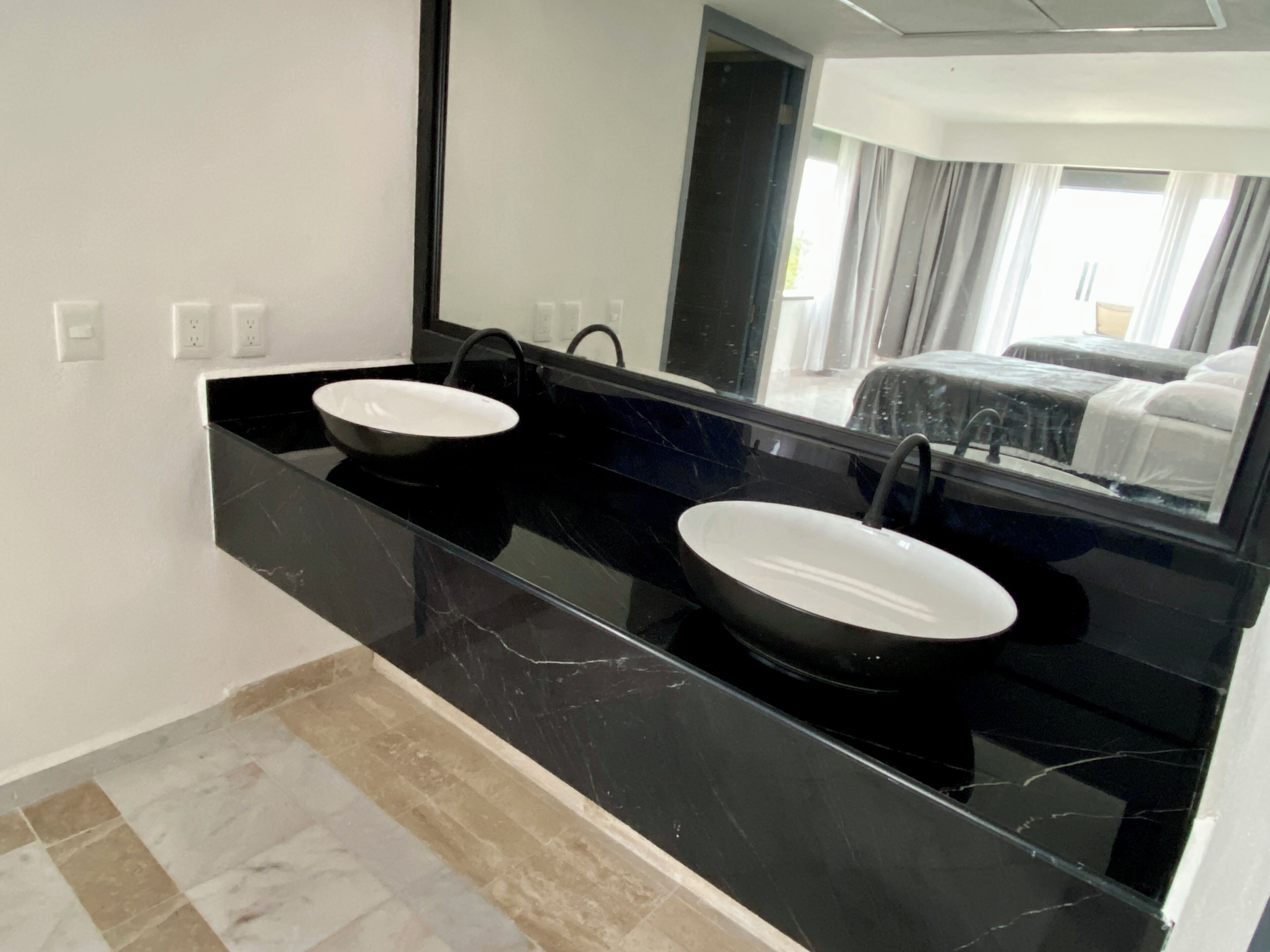 A modern bathroom with two sinks, a large mirror, and a view of a bedroom through the mirror.
