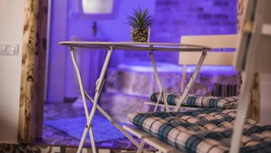 A room with a patterned floor, a small table with a potted plant, a chair with a checkered cushion, and a staircase in the background. - Old School (Ein Qiniyye)