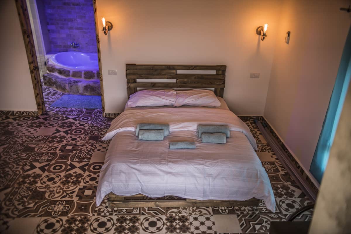 A bedroom with a wooden headboard, a patterned floor, and a view of a bathroom with a bathtub.