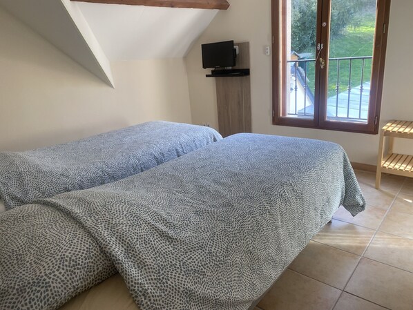 A bedroom with two beds covered in patterned bedding, a television on a wooden stand, and a view of a balcony with a railing. - Beauval Chambre (Seigy)