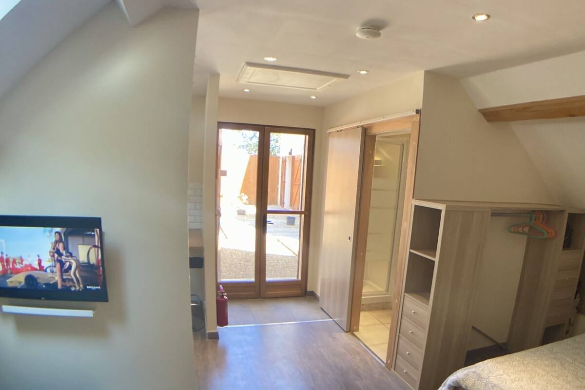 Habitación de hotel con un amplio ventanal corredero que da a un espacio exterior, una cama con cubrecama blanco, piso de madera y un televisor montado en la pared mostrando una escena de película.