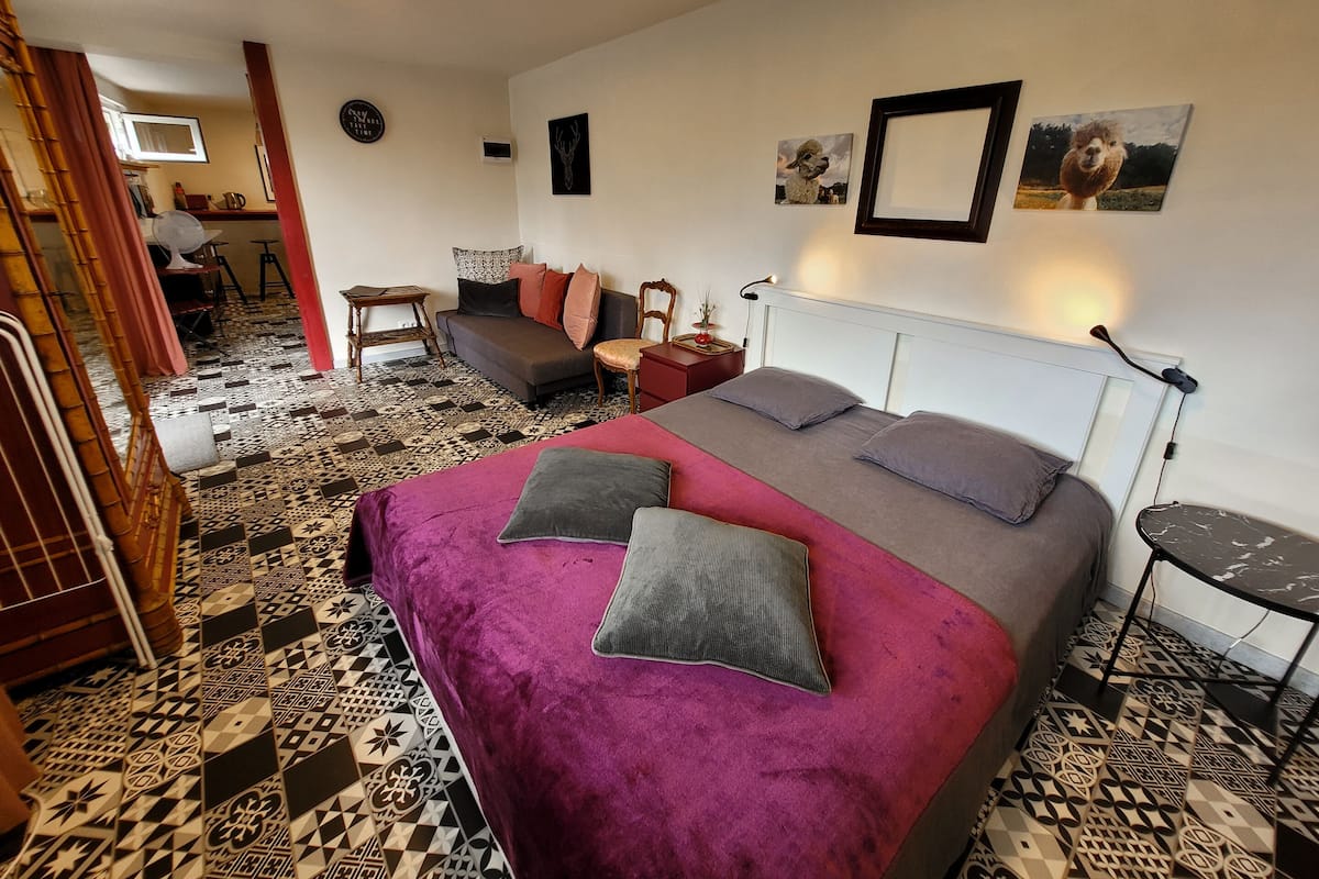 Une chambre avec un sol à motifs, un lit recouvert d’une couvre-lit violette, un canapé gris, une petite table avec une lampe et un téléviseur fixé au mur.