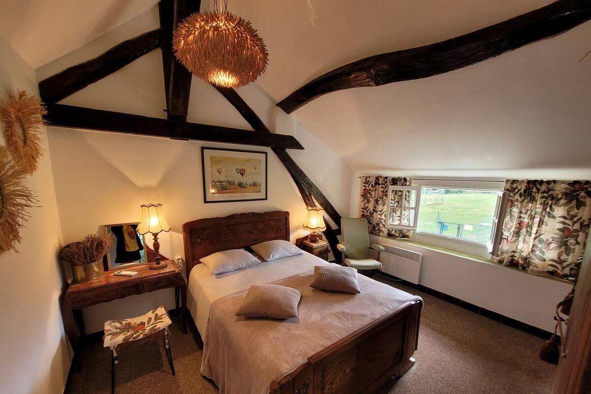 Une chambre à coucher avec un lit en bois, une table de chevet avec une lampe, une chaise, une fenêtre avec des rideaux et un tableau encadré accroché au mur.