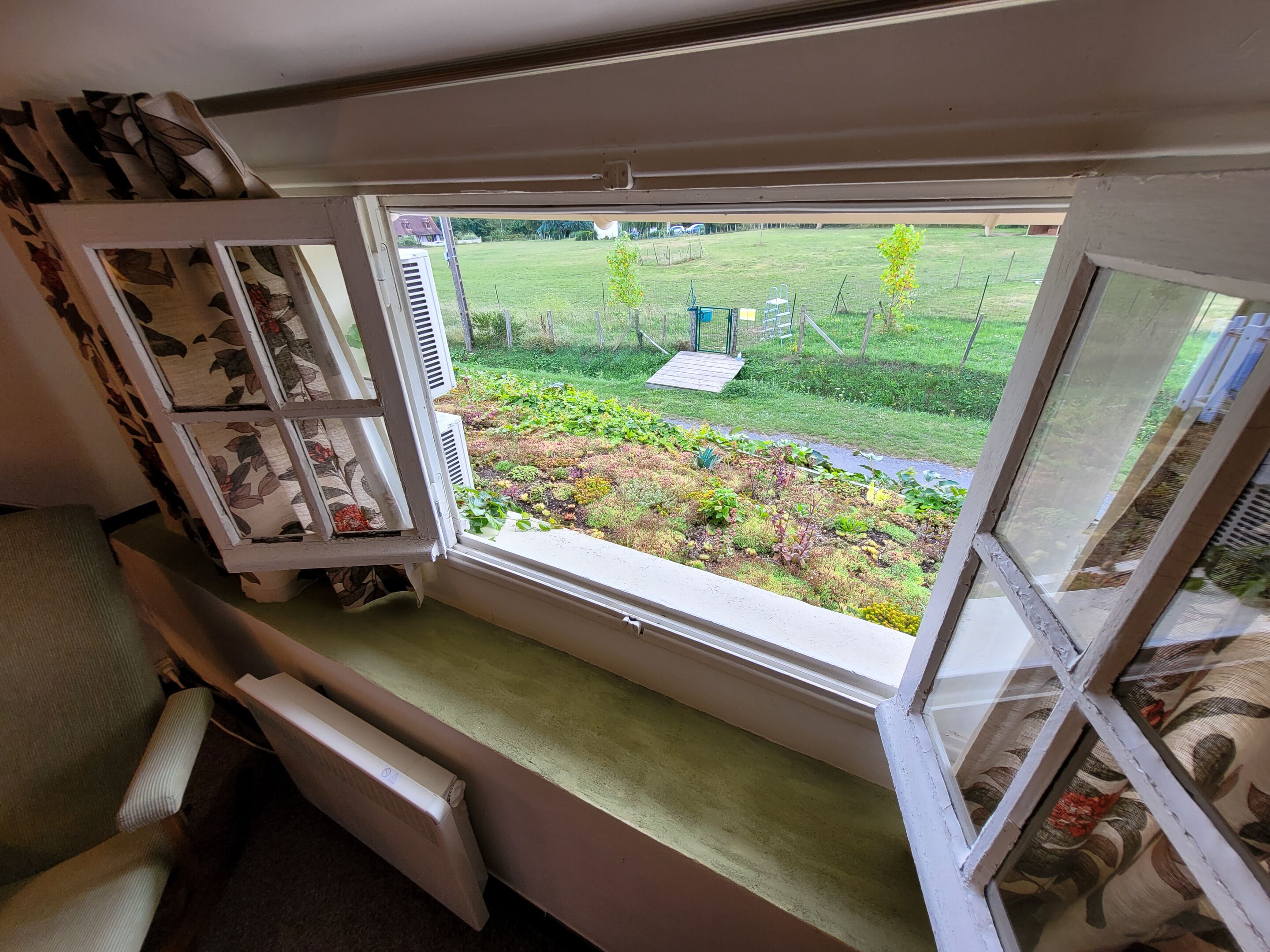 A room with a large window overlooking a garden. There is a radiator on the window sill and a chair in the foreground.