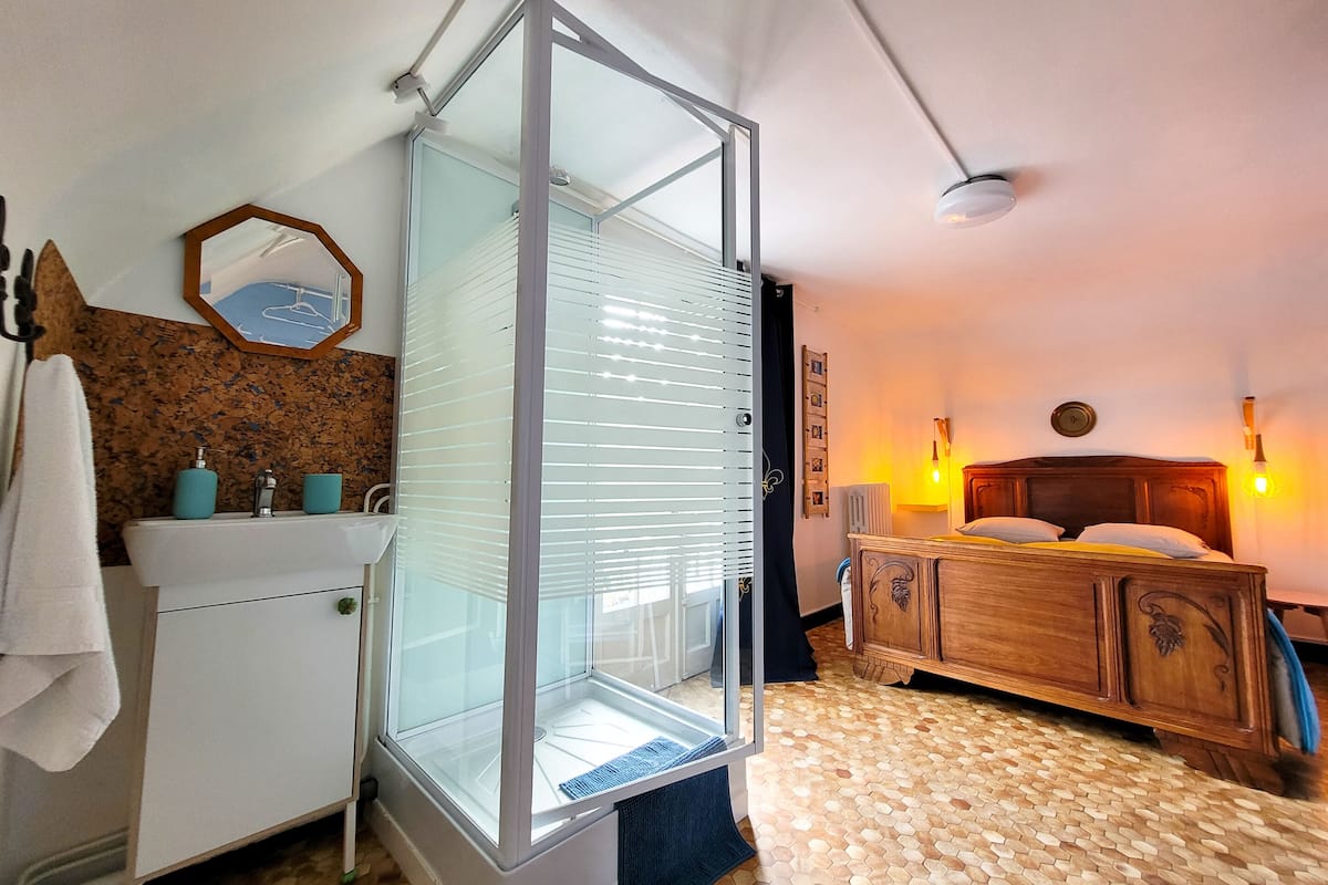 Une chambre d’hôtel dotée d’une douche en verre, d’un lit en bois, d’un petit lavabo blanc avec deux porte-savons bleus, d’une tête de lit en bois ornée d’une horloge et d’un sol à motifs hexagonaux.