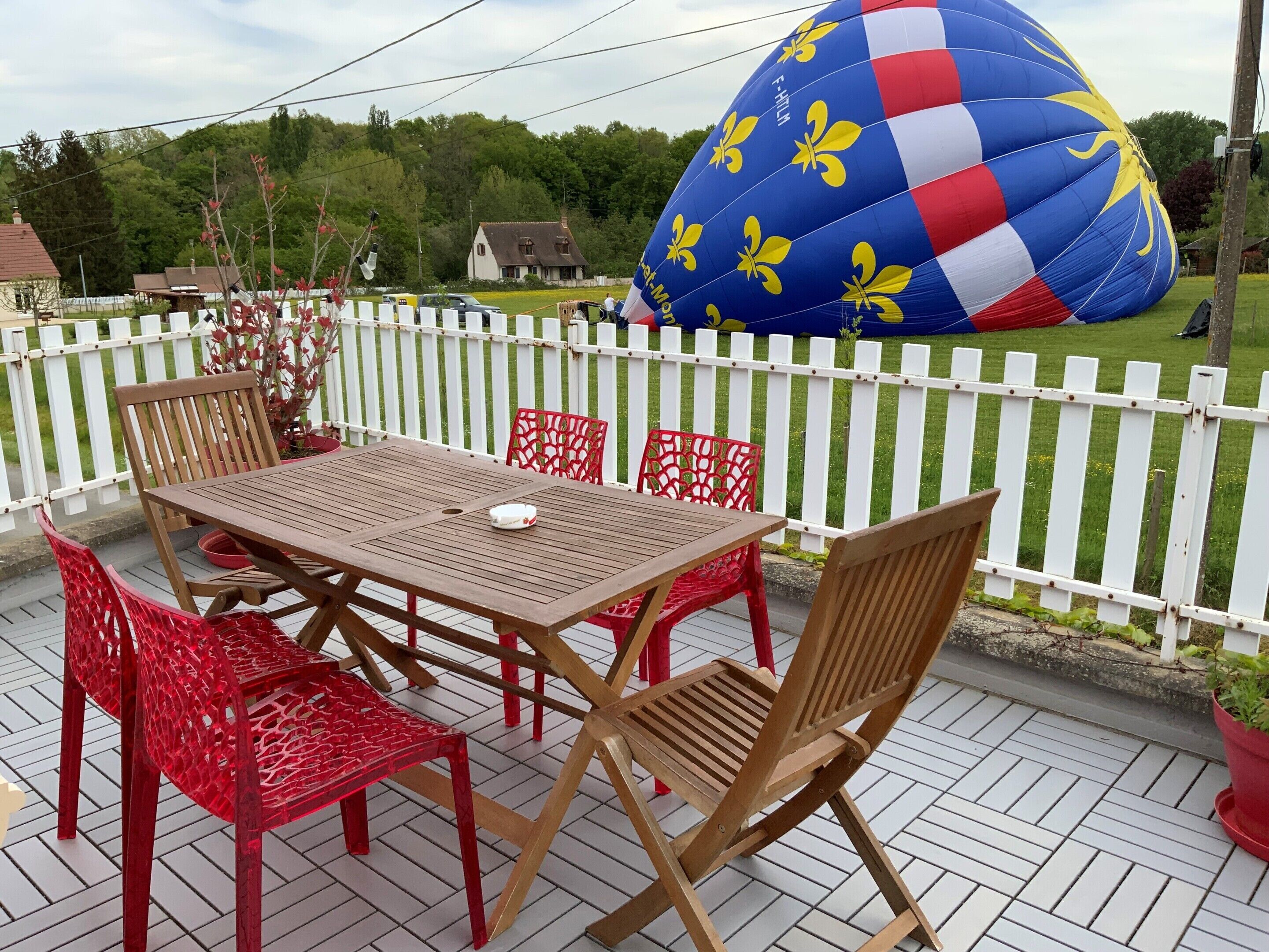 Eine Terrasse mit Holztisch und roten Stühlen, einem weißen Zaun und einer großen, blauen Heißluftballon im Hintergrund.