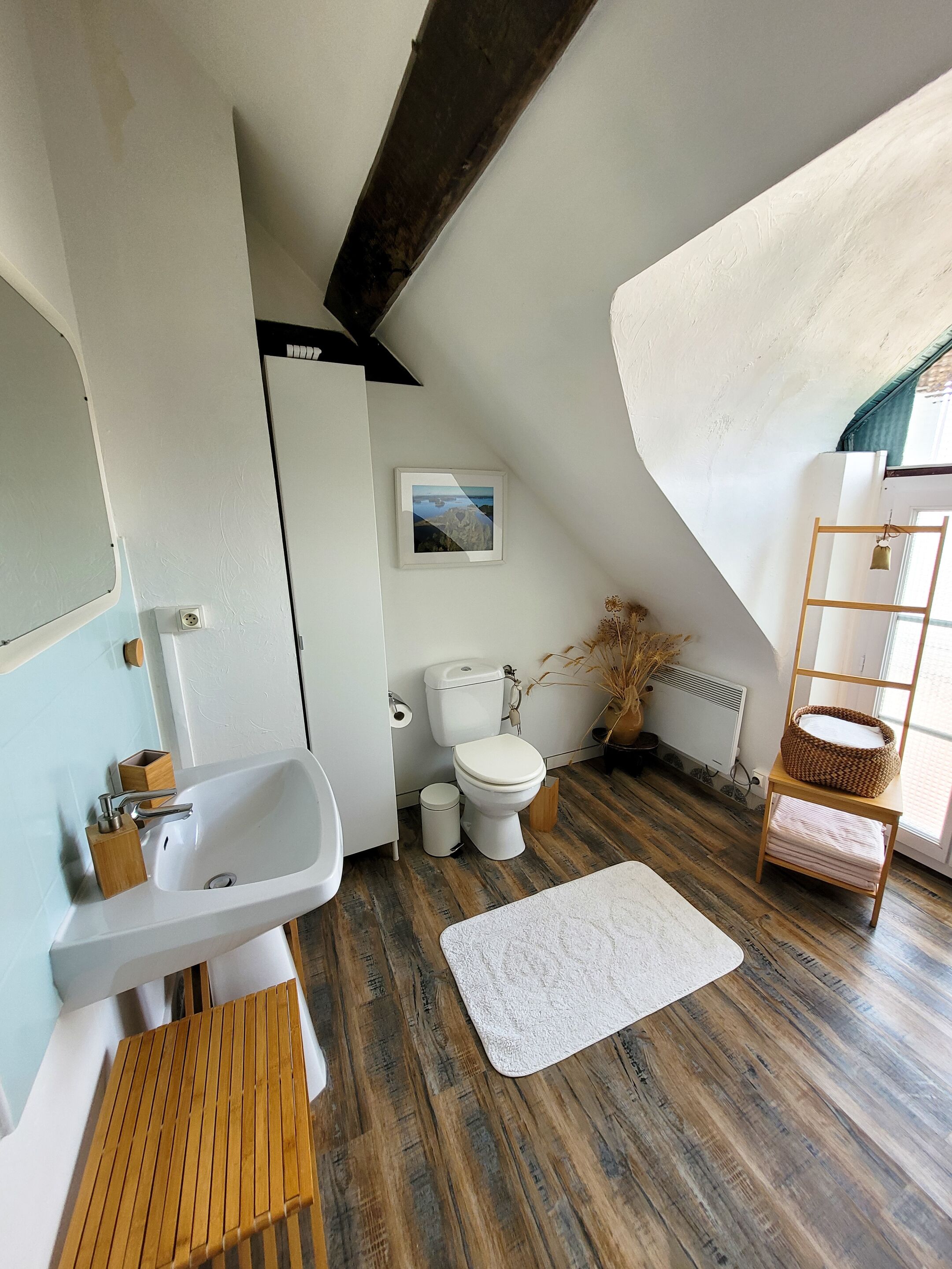 Un baño moderno con lavamanos, inodoro y bañera blancos. La habitación tiene suelo de madera y techo inclinado.