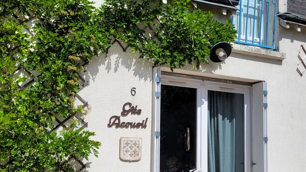 Un edificio con fachada blanca, un balcón azul y una puerta con panel de vidrio. Hay plantas en macetas y un letrero en la pared.