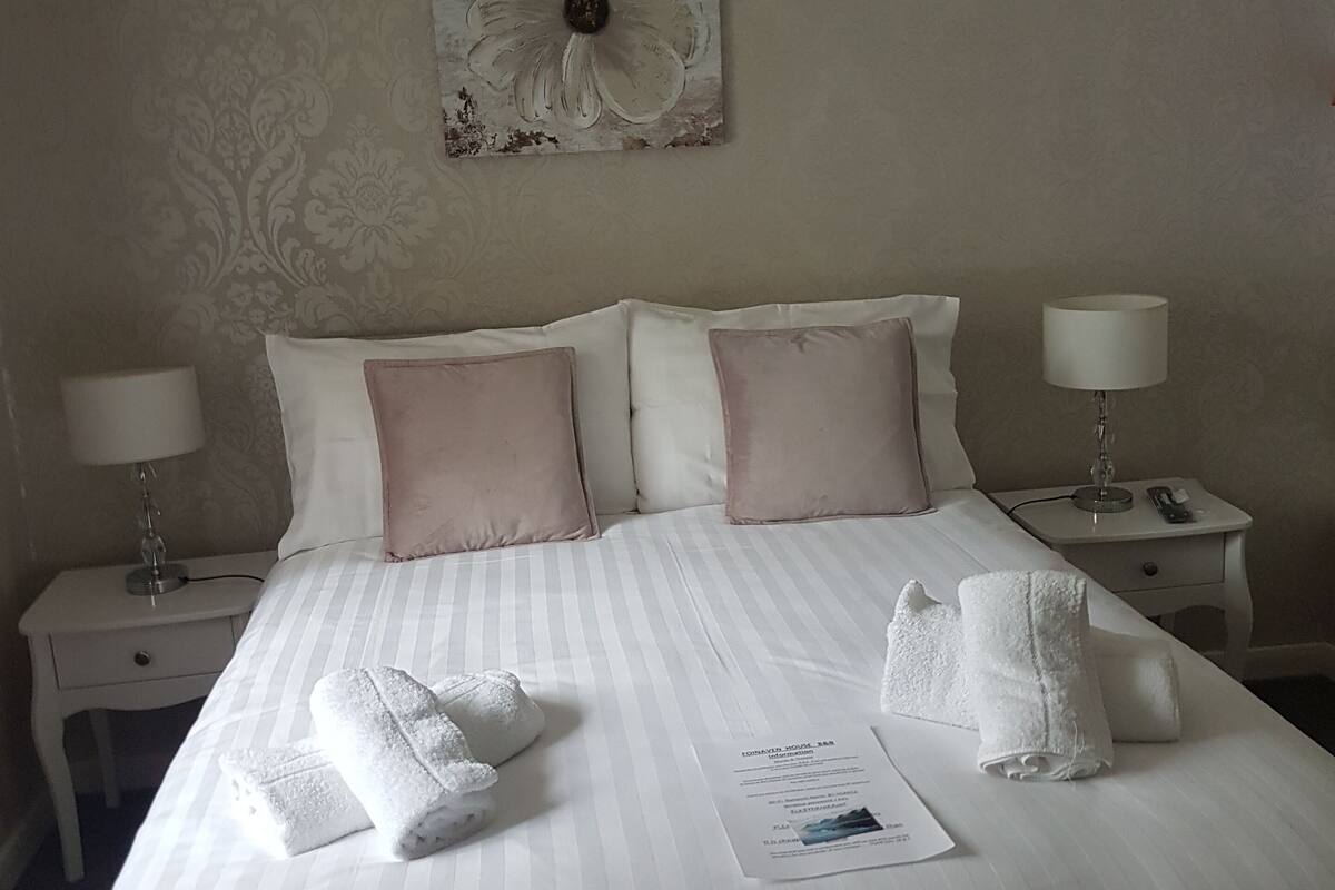A neatly made bed with white linens and pink pillows, a floral artwork on the wall, and a bedside table with a lamp and a phone.