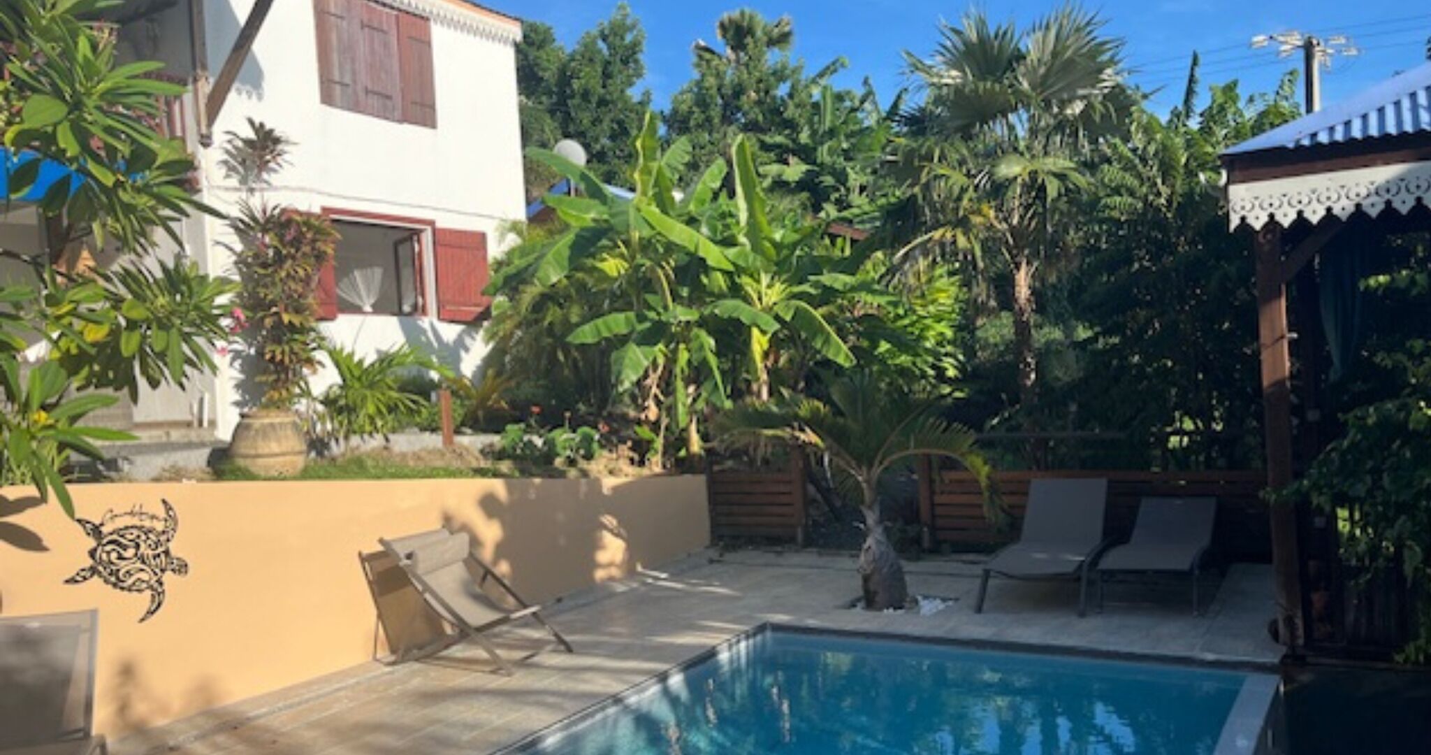 Una piscina con sillones de descanso, rodeada de plantas tropicales y un edificio blanco con contraventanas rojas.