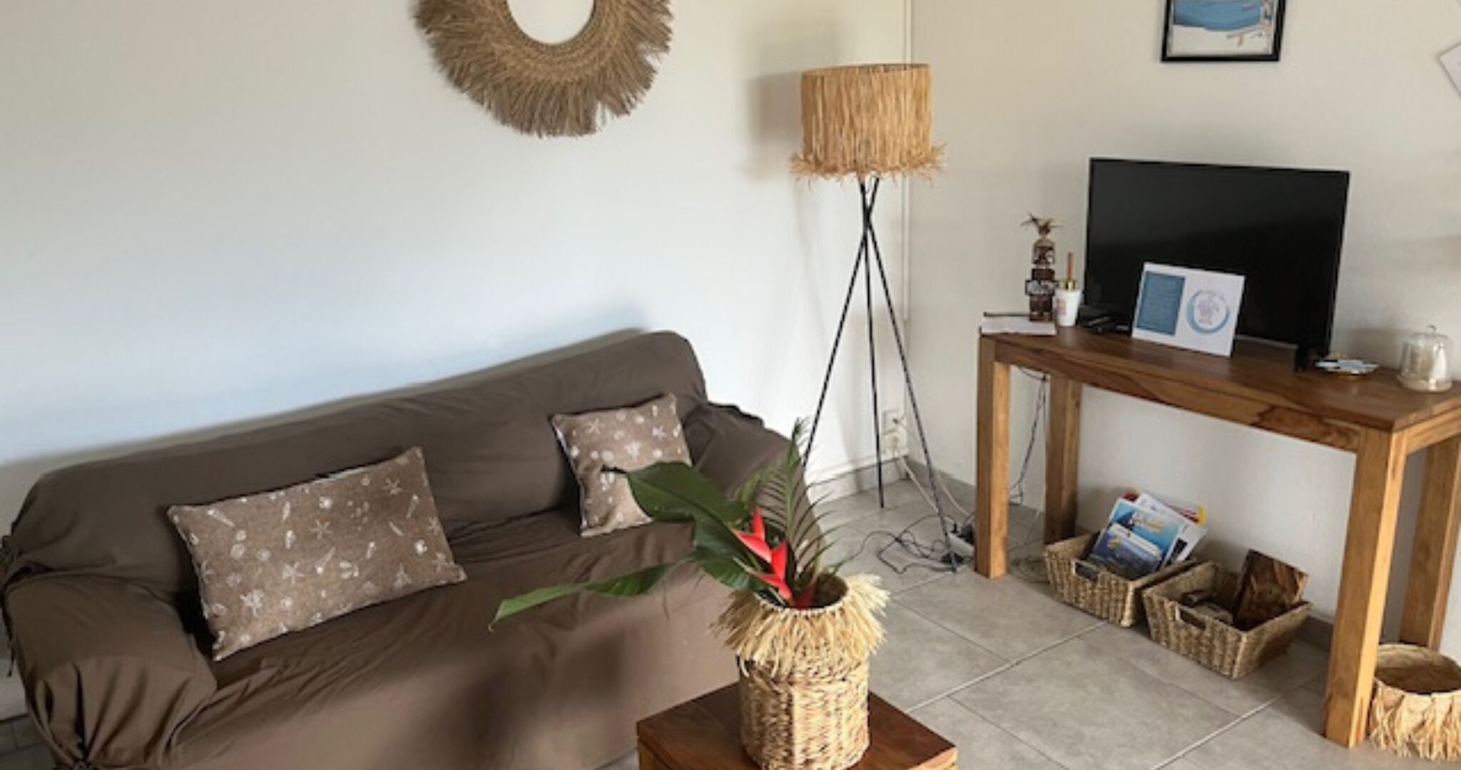 Una sala de estar con un sofá marrón, un soporte tejido para planta, una lámpara de pie, un mueble de televisión de madera y un cuadro en la pared.