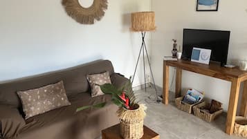 Una sala de estar con un sofá marrón, un soporte tejido para planta, una lámpara de pie, un mueble de televisión de madera y un cuadro en la pared.