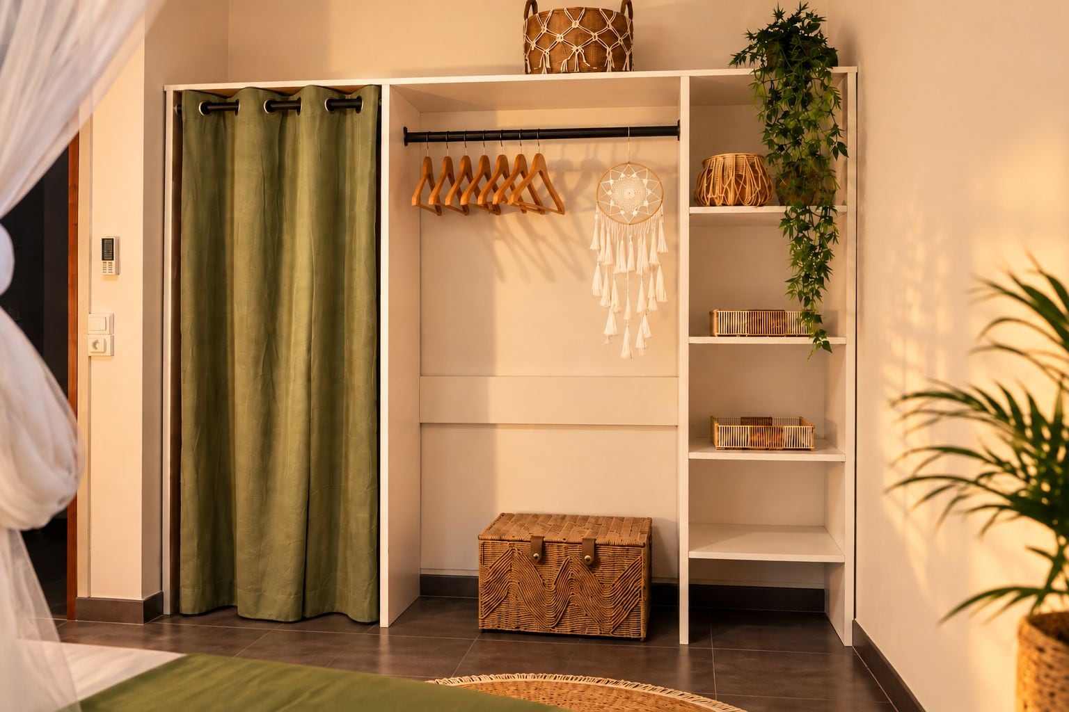 Ein Zimmer mit einem grünen Vorhang, einem Holz-Kleiderständer, einem Traumfänger, Korbwägelchen und einer Topfpflanze.