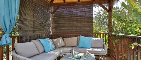 A covered outdoor seating area with a wicker sofa and a glass coffee table, surrounded by wooden railings and lush greenery.