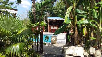Uma área de piscina com deck de madeira, cercada por plantas tropicais e um edifício com telhado azul.