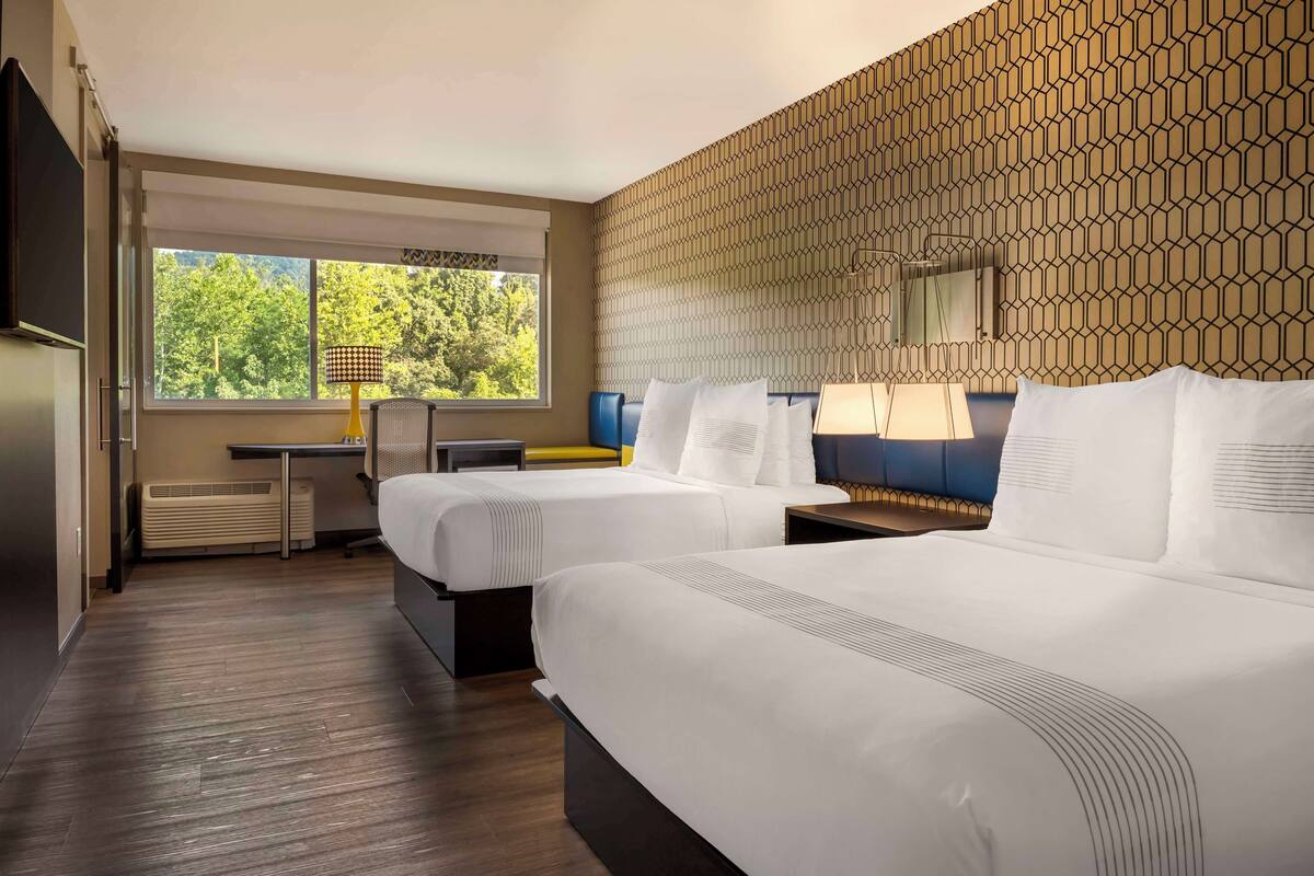 A hotel room with two beds, a desk, a chair, a lamp, and a view of trees through the window.