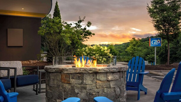 An outdoor seating area with a stone fire feature, blue Adirondack chairs, and a view of a lush landscape with a sunset. - GLō Hotel Asheville-Blue Ridge Parkway (Asheville)