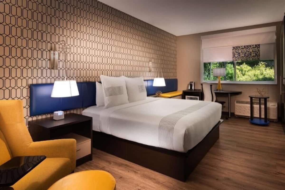 A modern hotel room with a large bed, a desk, a chair, and a window with a view.