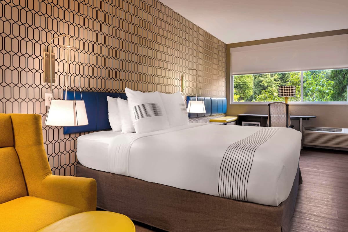 A hotel room with a patterned wall, a large bed with white bedding, a yellow armchair, a desk with a lamp, and a view of greenery through the window.
