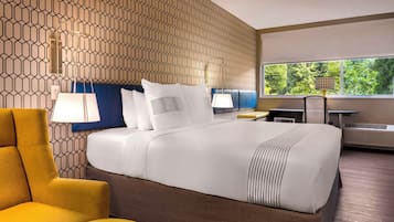 A hotel room with a patterned wall, a large bed with white bedding, a yellow armchair, a desk with a lamp, and a view of greenery through the window.