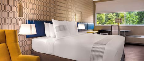 A hotel room with a patterned wall, a large bed with white bedding, a yellow armchair, a desk with a lamp, and a view of greenery through the window.