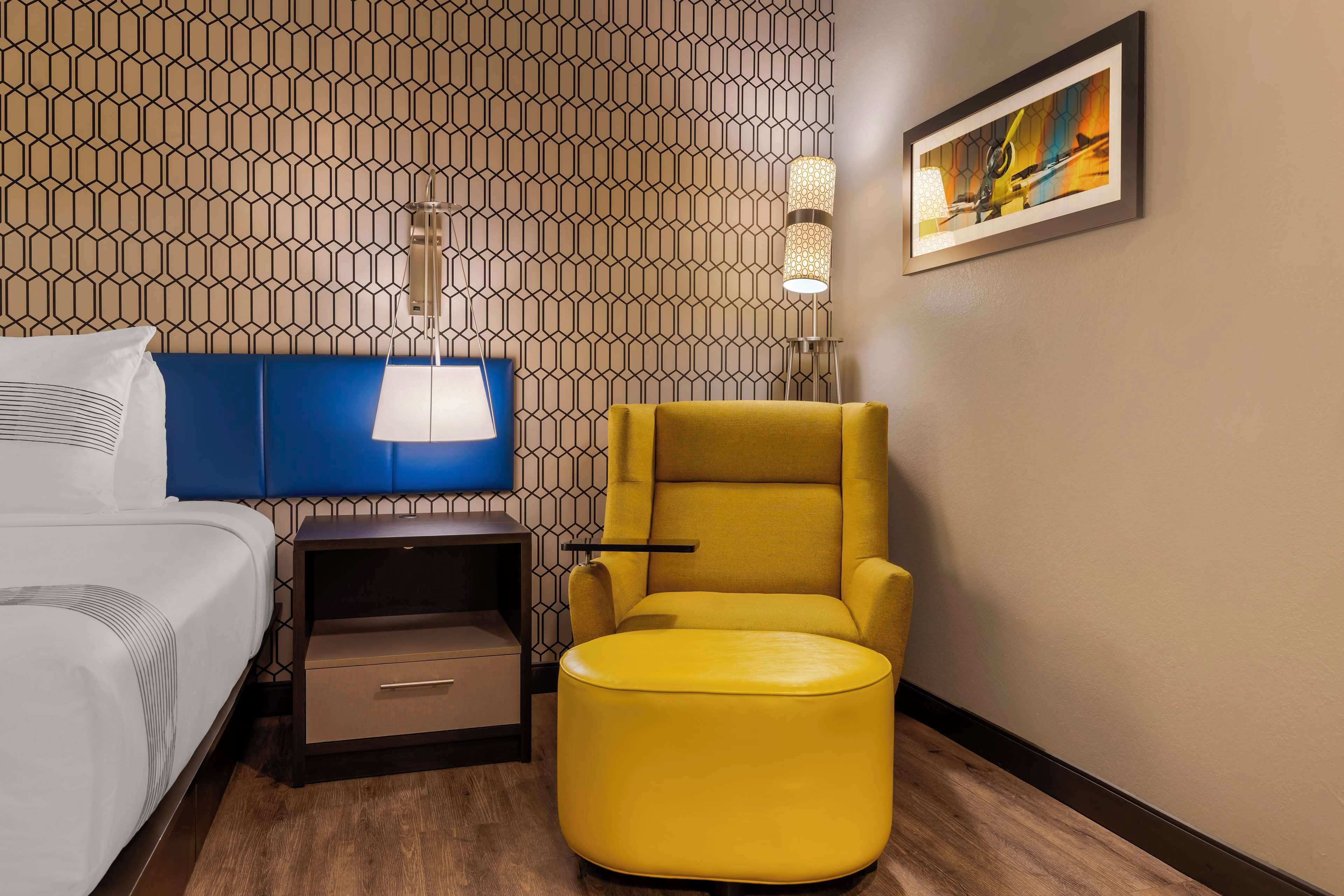 Una habitación de hotel con cama, cabecera azul, sillón amarillo, mesita de noche, lámpara y un cuadro en la pared.