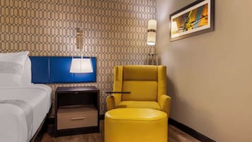 Una habitación de hotel con cama, cabecera azul, sillón amarillo, mesita de noche, lámpara y un cuadro en la pared.