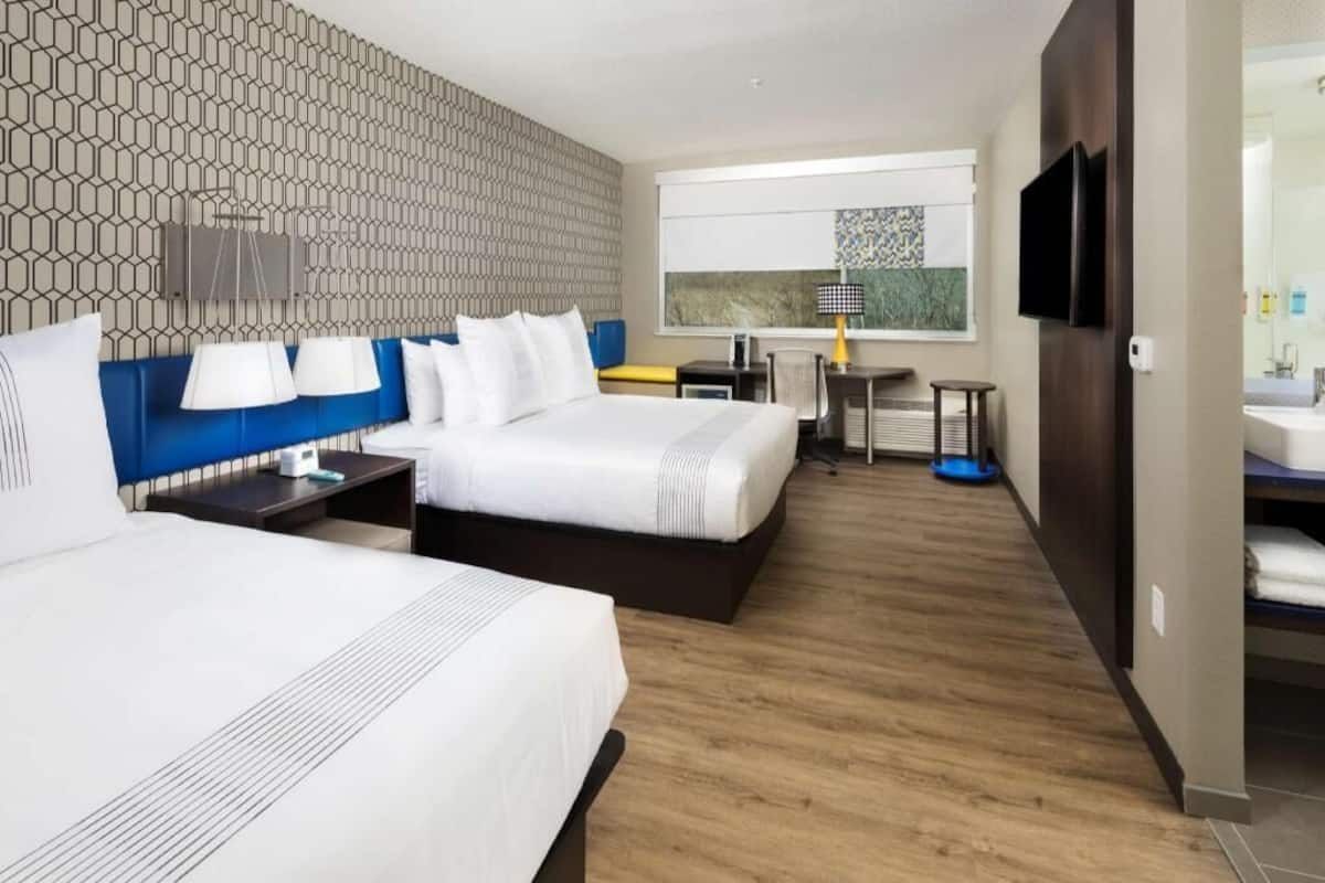 A hotel room with two beds, a desk, a chair, a TV, and a bathroom with a sink and mirror.