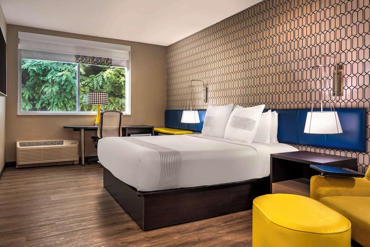 A hotel room with a large bed, a desk, and a chair. There is a window with a view of greenery outside.