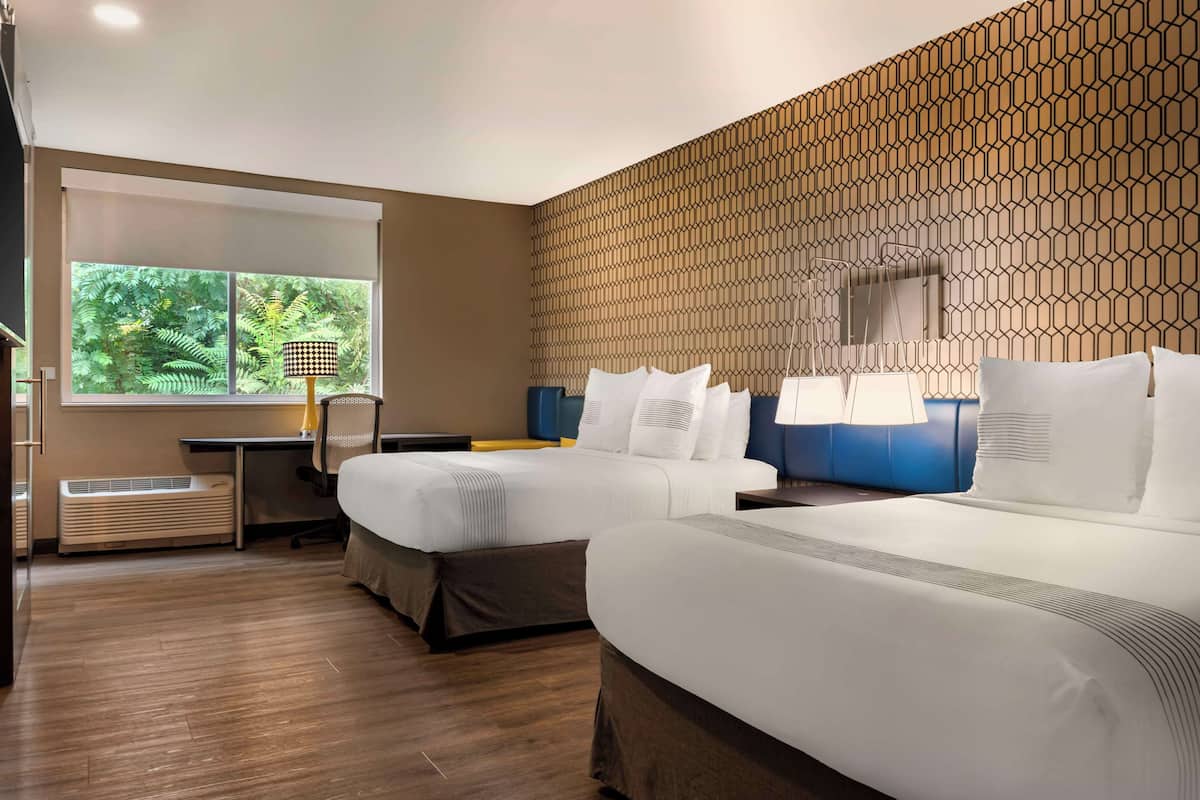 A hotel room with two beds, a large window with a view of greenery, a wall with a geometric pattern, and a desk with a lamp.