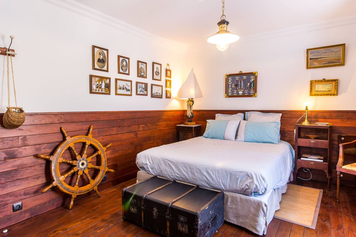 A bedroom with a wooden headboard, a large bed with white bedding, a ship's wheel decoration, and several framed pictures on the wall.