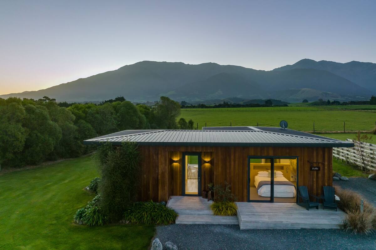 A modern cabin with a large window, a small porch, and a view of mountains.