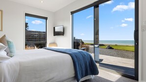 A bedroom with a large bed, a TV mounted on the wall, and a sliding glass door leading to a balcony with a view of the beach. - Glenburn Coastal Retreat (Hapuku)