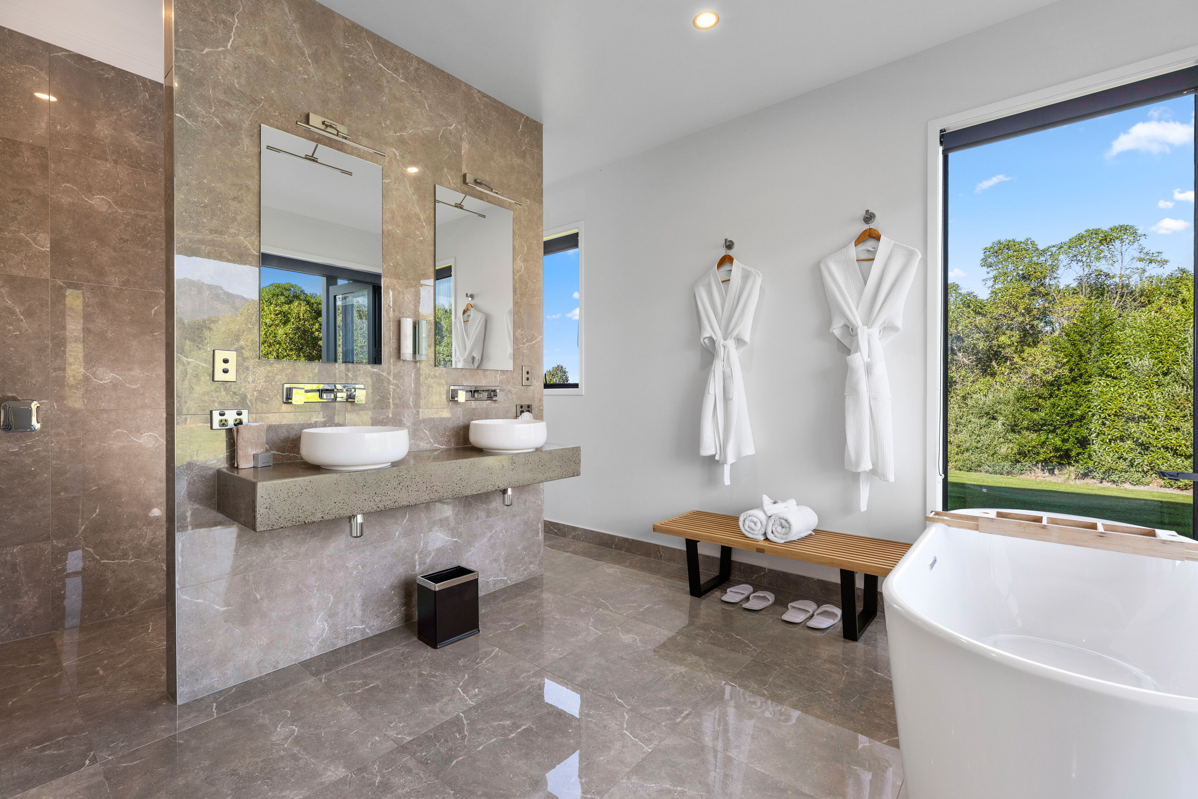 A modern bathroom with two sinks, a large mirror, a bench, and a bathtub.