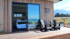 A modern wooden building with a sliding glass door leading to a bedroom with a bed and a view of the mountains. There are two Adirondack chairs on the deck. - Glenburn Coastal Retreat (Hapuku)